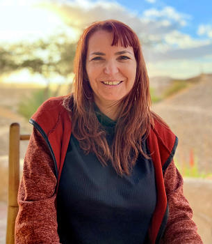 A woman in a red vest with red hair sits under a pergola and smiles at the camera.