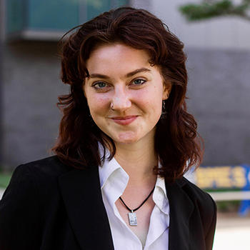 A young woman in a dark jacket with dark hair and a white shirt smiles at the camera.